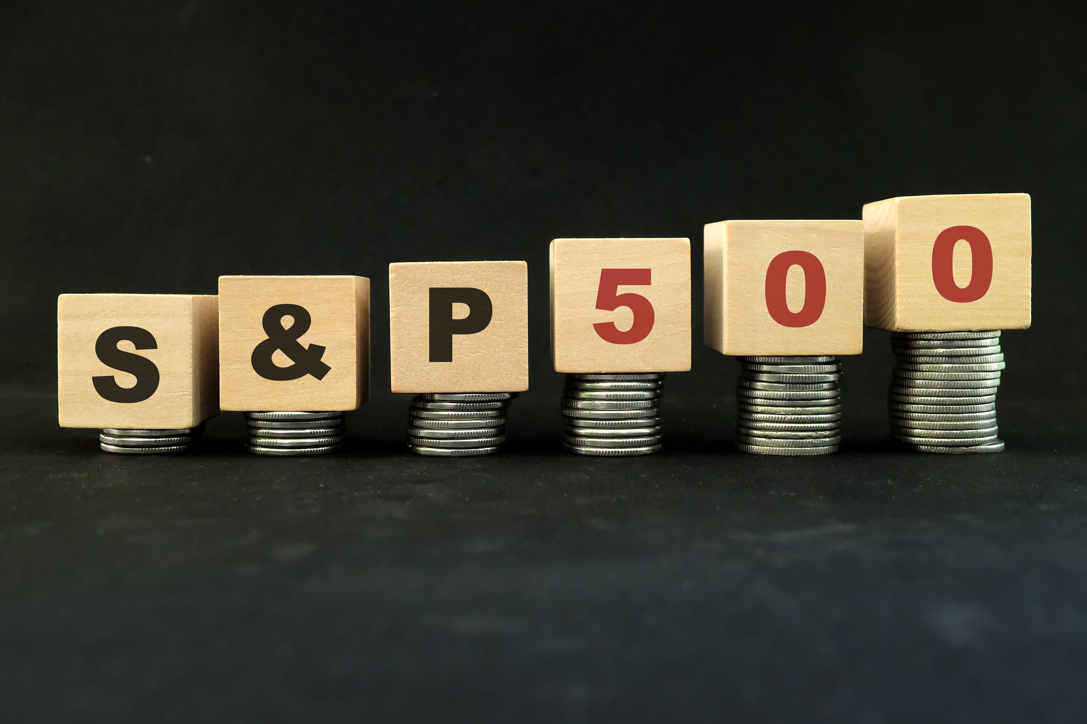 Wooden blocks lettered S&P 500, each on a taller stack of coins than the last, against a black background.